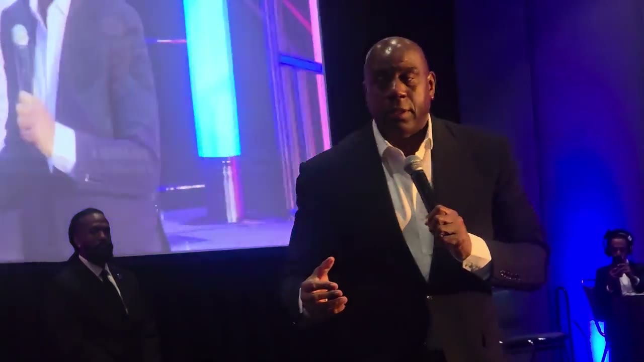 A speaker with a microphone addresses a conference audience in front of a blue-lit stage screen